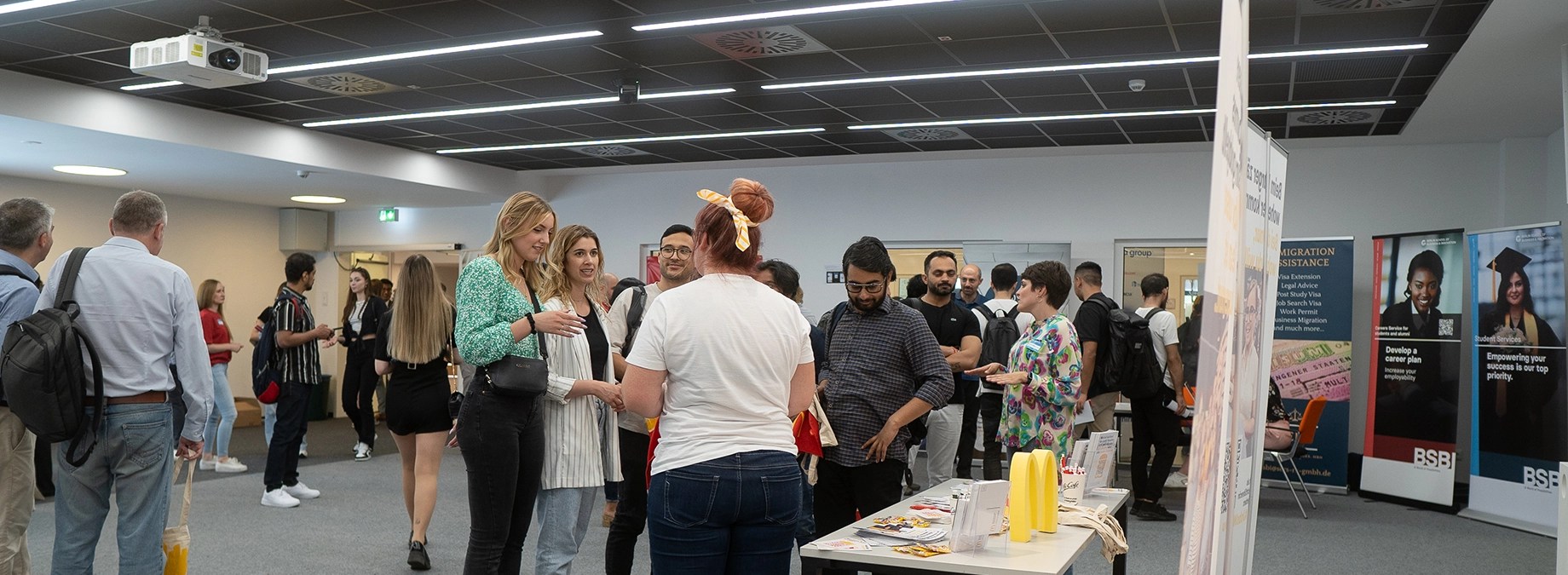 Studierende unterhalten sich mit Unternehmensvertreterinnen an Informationsständen bei einer Karrieremesse an der University of Europe for Applied Sciences. Im Hintergrund sind Roll-Ups mit Informationen zu Karriereservices und Einwanderungshilfe sichtbar